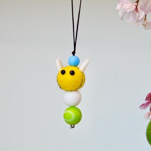 May include: A handmade bee-themed ornament. The bee has a yellow body, white wings, and black eyes, with a blue bead on top. Below the bee is a white bead and a green baseball-style bead. The ornament is suspended from a black string.