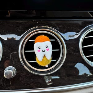 May include: A felt tooth-shaped car air freshener. The tooth has a white enamel, a yellow root, and an orange cap. It features a smiling face with pink heart-shaped cheeks. The air freshener is placed in a car's air vent.
