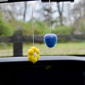 May include: Two car air fresheners suspended from a car's rearview mirror. One is a yellow flower shape with a white centre, and the other is a blue heart shape. Both are made of a fuzzy material and hang by thin strings.
