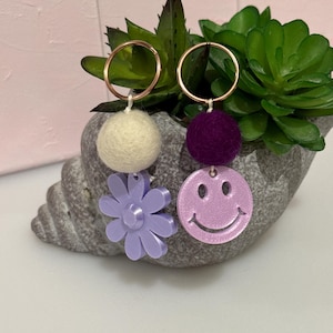 May include: Two keychains featuring gold-tone metal rings. One keychain has a cream-coloured felt ball and a light purple flower charm. The other keychain has a dark purple felt ball and a light purple circular charm with a smiley face embossed on it.