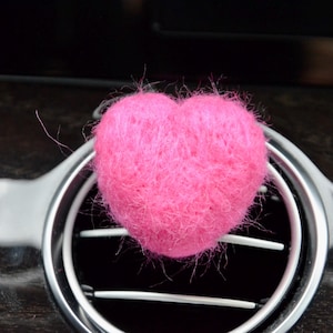 May include: A bright pink, fuzzy heart-shaped car air freshener. The heart is positioned in front of a car's air vent, with a dark wood-grain dashboard in the background. The air vent is silver and black.
