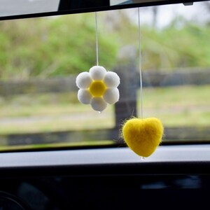May include: Two car air fresheners suspended by string. One is a white daisy with a yellow centre, and the other is a yellow heart. The background is a blurred view of a green landscape.