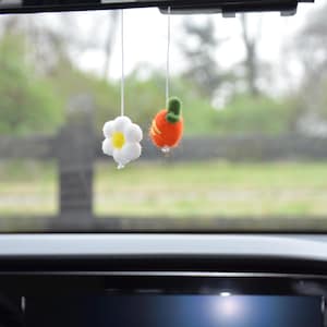May include: Two hanging car charms: a white daisy with a yellow centre and an orange carrot with a green stem. Both charms are suspended by thin white strings, with a blurred outdoor background visible.