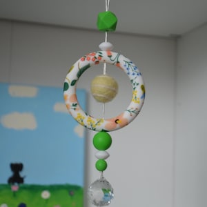 May include: A hanging baby mobile with a floral patterned ring, a yellow felt ball, and green and white beads. A clear crystal ball hangs at the bottom. The mobile is suspended from a white string.