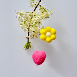 May include: A branch with white blossom and two hanging felt decorations. A bright yellow flower with a white centre and a pink heart are suspended from the branch with white string. The background is a light grey colour.