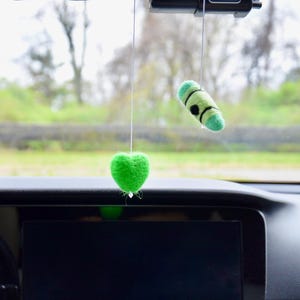 May include: Two handmade car air fresheners hanging from a car's rearview mirror. One is a bright green heart shape, and the other is a green cylinder with black accents. The background shows a blurred view of trees and a road.