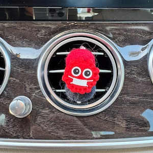 May include: A red, plush air vent charm shaped like a cartoon virus wearing a white face mask. The charm has large, black eyes and is attached to a gray, fuzzy base. The charm is placed in a car's air vent.
