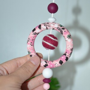 May include: A hanging mobile featuring a pink floral patterned ring, a burgundy felt ball, and assorted beads. The mobile is suspended by a white string and has a large crystal at the bottom. The ring is approximately 10 cm in diameter.