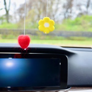 May include: A car interior featuring a black dashboard and a large screen. Suspended from above are two decorative ornaments: a red heart and a yellow flower with a white centre. The background shows a blurred view of greenery.