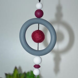 May include: A hanging mobile featuring a gray ring, white and burgundy beads, and a red felt ball. The mobile is suspended by a black string, creating a modern and minimalist design. The color palette includes gray, white, and burgundy.