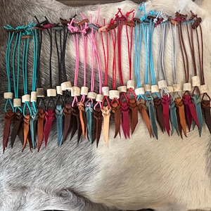 May include: A collection of leather cord zip pulls featuring various colours including turquoise, black, pink, red, blue, grey, and brown. Each pull is accented with a small, light-coloured cylindrical bead and finished with a knotted leather end.