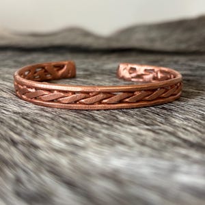 May include: A copper cuff bracelet featuring a central braided design flanked by smooth, solid bands. The metal has a warm, reddish-brown metallic finish with a visible texture throughout the surface. The bracelet is open-ended, showing a repeating cutout pattern along the inner edges of the band.