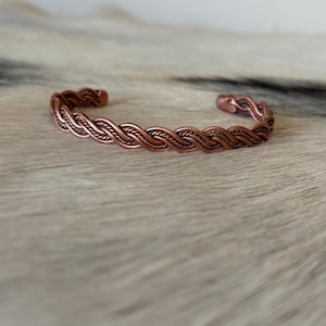 May include: A copper-colored cuff bracelet featuring a continuous braided or twisted wire design. The metal has a warm, reddish-brown metallic finish with a repeating pattern of intertwined strands that form a circular, open-ended band structure.