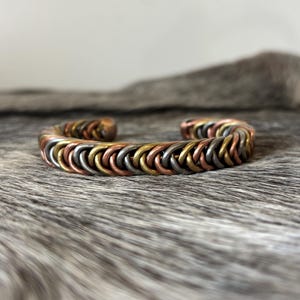 May include: A cuff bracelet constructed from interlocking metal rings in alternating shades of copper, brass, and silver. The circular links are woven together in a repeating pattern to form a continuous band with an open gap at the back.