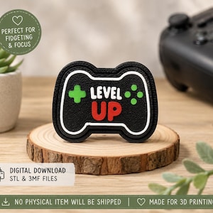 May include: A black, white, green, and red 3D printed object shaped like a video game controller. The center features the text "LEVEL UP" in white and red lettering, flanked by a green plus sign and four green dots on a textured black surface.