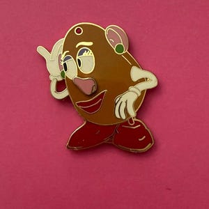 May include: A Mr. Potato Head enamel pin with a gold outline. The pin features a brown potato-shaped body, red shoes, and white gloved hands. The face has a pink nose, a red smiling mouth, and green and white eyes. The background is a solid pink.