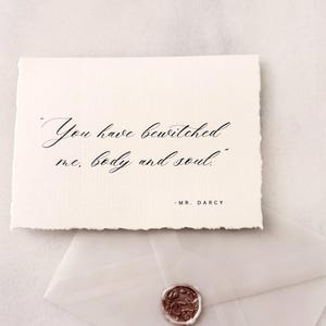 May include: A white card with the quote "You have bewitched me, body and soul" in elegant calligraphy, resting above a translucent envelope sealed with a brown wax stamp. The quote is attributed to Mr. Darcy.