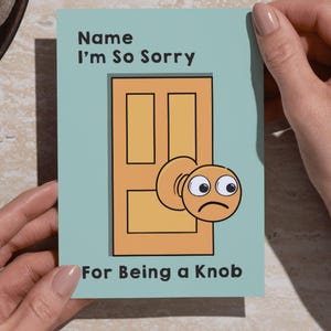 May include: A greeting card with a light blue background and the text "Name I'm So Sorry For Being a Knob." The card features a cartoon door with a sad doorknob. The card is held by two hands.
