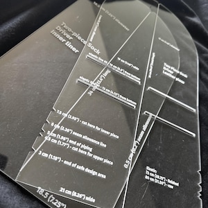 May include: Two clear acrylic templates for a two-piece sock driver. The templates feature white printed text and markings, including measurements in centimeters and inches, such as 18.5 cm (7.25") width, 34 cm (13.4") length, and various seam allowance lines.