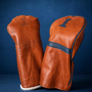 May include: Two tan leather golf club head covers featuring the number 1. The left cover has a debossed number 1 and a white faux shearling lining at the base. The right cover features a dark blue leather stripe and a dark blue number 1 patch.