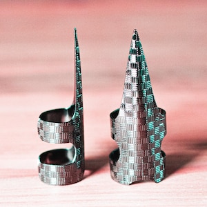 Pode incluir: Dois protetores de dedos prateados com um padrão tecido. Um é alto e esguio, o outro tem formato de cone. Ambos têm uma faixa curva para o dedo e uma superfície texturizada. O fundo é rosa suave.