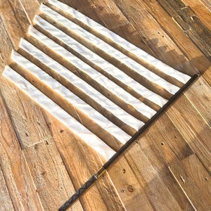 May include: A white and gold fan with a dark brown handle. The fan has a series of white, rectangular panels. The handle is wrapped in a dark material with a gold accent at the base. The fan is lying on a wooden surface.