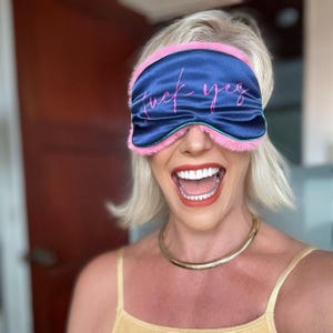 Puede incluir: Un antifaz de dormir de satén azul oscuro con ribete de piel sintética rosa y el texto "fuck yes" escrito en cursiva rosa. El antifaz presenta una forma contorneada para cubrir los ojos, con una textura suave y afelpada en los bordes.