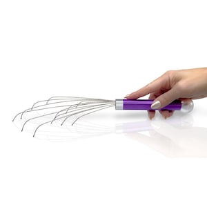 Può includere: Un massaggiatore per cuoio capelluto con un manico cilindrico viola brillante e un tappo terminale sferico bianco liscio. Il dispositivo include molteplici punte in filo metallico sottili e flessibili che si estendono da una base argentata.