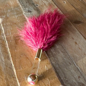 Könnte beinhalten: Ein rosa Federstaubwedel mit einem goldenen und transparenten Griff. Der Staubwedel hat eine große Ansammlung von leuchtend rosa Federn. Der Griff hat eine goldfarbene Metallspitze und einen transparenten, zylindrischen Körper.