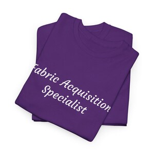 May include: Two folded purple t-shirts with the text "Fabric Acquisition Specialist" in white script. The shirts are made of a soft material and are neatly stacked on a white background.