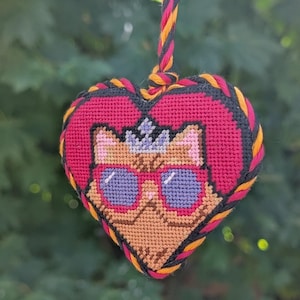 Op de afbeelding: Een hartvormig kruissteek ornament met een beige kat die een paarse zonnebril en een kleine blauwe kroon draagt. De kat is gecentreerd op een rode achtergrond en het hart is afgewerkt met een gedraaid zwart, oranje, roze en zwart koord.