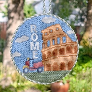 Op de afbeelding: Een rond borduurornament met een afbeelding van het Colosseum in Rome, een rode scooter en het woord ROME in witte letters tegen een blauwe lucht met witte wolken. Het ronde stuk is afgewerkt met een gedraaide koordrand.