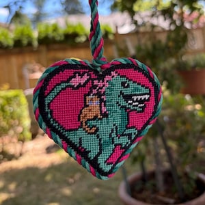 May include: A heart-shaped needlepoint ornament featuring a green dinosaur with a small brown creature on its back, set against a bright pink background. The piece is framed by a thick black border and finished with a twisted green and pink rope.