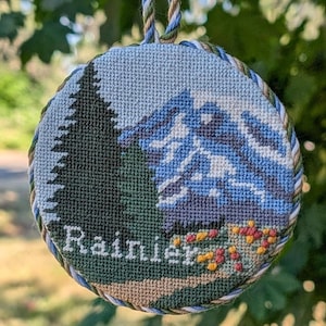 May include: A round cross-stitch ornament featuring a mountain landscape with a dark green pine tree, blue and white snow-capped peaks, and small yellow and red flowers. The word Rainier is stitched in white at the bottom with a twisted cord edge.