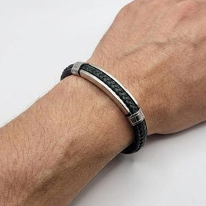May include: Black braided leather bracelet with a silver-tone metal centre bar and accents. The bracelet is worn on a wrist and has a classic, masculine design. The bracelet is approximately 20 cm in length.