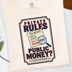 May include: A cream-colored t-shirt featuring a graphic design with the text PRIVATE RULES PUBLIC MONEY? and a tax receipt illustration with a magnifying glass over the word ACCOUNTABILITY. The shirt has a Comfort Colors brand label at the neck.