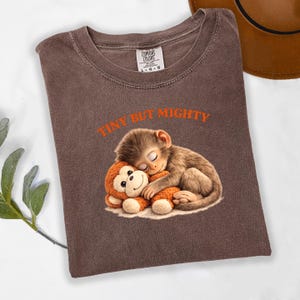 May include: Brown t-shirt with the text "TINY BUT MIGHTY" in orange. The shirt features a graphic of a sleeping monkey hugging a stuffed monkey. The shirt is a Comfort Colors brand.