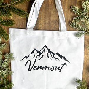 Può includere: Borsa tote in tela bianca con grafica di montagna nera e la scritta "Vermont". La borsa ha due manici bianchi ed è circondata da rami di pino verdi.