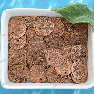 May include: A white ceramic dish filled with copper-colored metal charms. The charms are round and have a decorative, floral design.