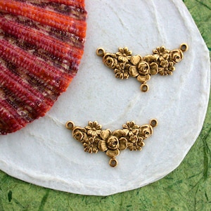 May include: Two antique gold-colored floral connectors. Each connector features a detailed arrangement of roses and other flowers. The connectors are displayed on a white surface with a red and orange seashell and a green textured background.