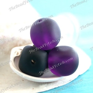 May include: Three glass beads in shades of purple and black rest in a white shell-shaped dish. Two round, frosted purple beads and one square, matte black bead are visible. The background is a light blue.