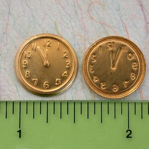 May include: Two gold-coloured clock faces with black hands and Roman numeral markings. The clock faces are approximately 1 inch in diameter and are sitting on a green ruler.