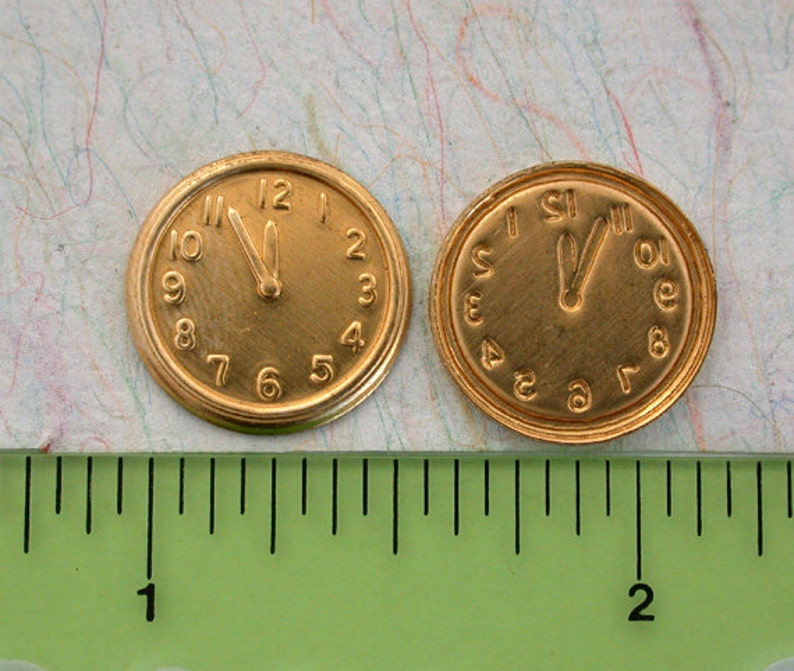 May include: Two gold-coloured clock faces with black hands and Roman numeral markings. The clock faces are approximately 1 inch in diameter and are sitting on a green ruler.