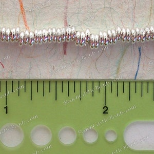 May include: Close-up of a strand of shiny, silver-colored beads. The beads are small, round, and evenly spaced. A green ruler with inch markings is in the foreground, measuring the beads. The background is a textured, off-white surface with colorful lines.