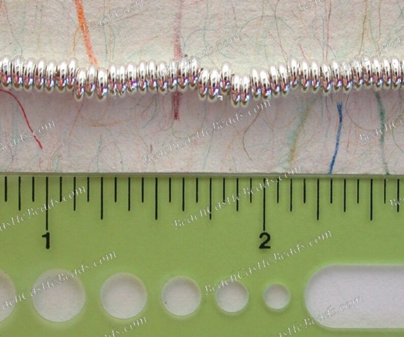 May include: Close-up of a strand of shiny, silver-colored beads. The beads are small, round, and evenly spaced. A green ruler with inch markings is in the foreground, measuring the beads. The background is a textured, off-white surface with colorful lines.