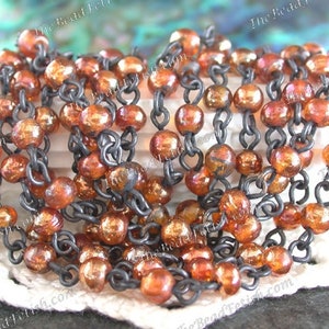 May include: A close-up of a chain of small, iridescent copper-colored beads strung on a black metal chain. The beads have a slightly irregular shape and a shiny, metallic finish.