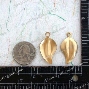 K&ouml;nnte beinhalten: Zwei goldfarbene, blattf&ouml;rmige Metallanh&auml;nger mit einem Loch oben zum Aufh&auml;ngen. Die Anh&auml;nger sind etwa 2,5 cm lang und werden neben einem US-amerikanischen Vierteldollar zur Gr&ouml;&szlig;enreferenz gezeigt.