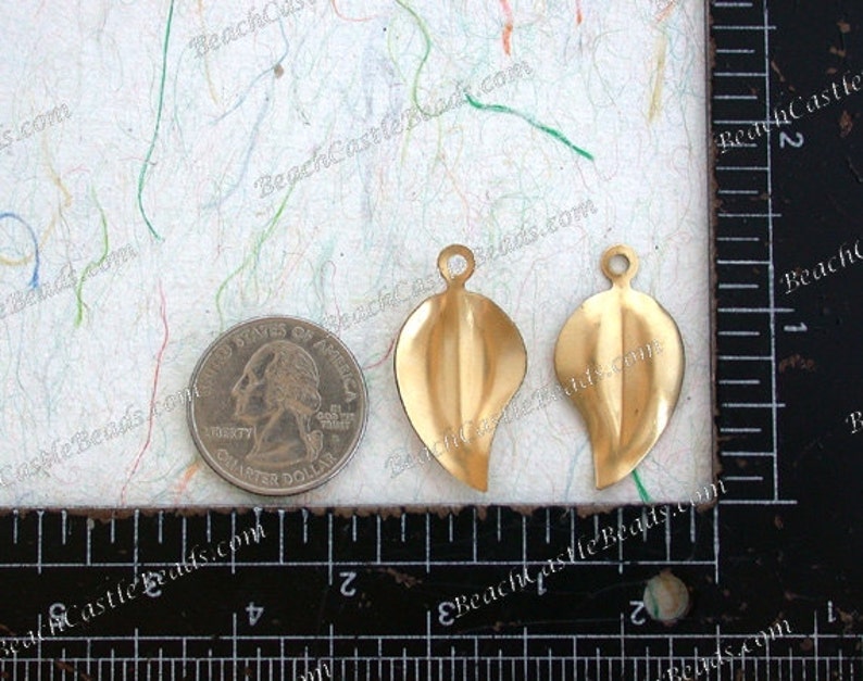 K&ouml;nnte beinhalten: Zwei goldfarbene, blattf&ouml;rmige Metallanh&auml;nger mit einem Loch oben zum Aufh&auml;ngen. Die Anh&auml;nger sind etwa 2,5 cm lang und werden neben einem US-amerikanischen Vierteldollar zur Gr&ouml;&szlig;enreferenz gezeigt.