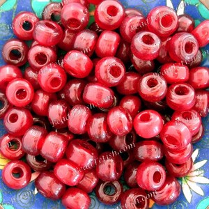 May include: A collection of translucent red glass beads with large holes, filling a decorative blue and green bowl. The beads are cylindrical and appear to be strung together. The bowl has a floral design around the rim.