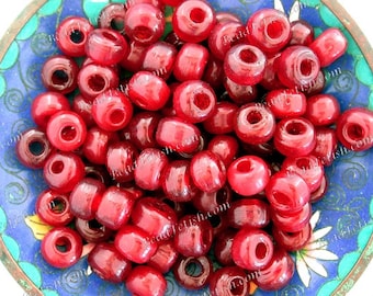 May include: A collection of translucent red glass beads with large holes, filling a decorative blue and green bowl. The beads are cylindrical and appear to be strung together. The bowl has a floral design around the rim.
